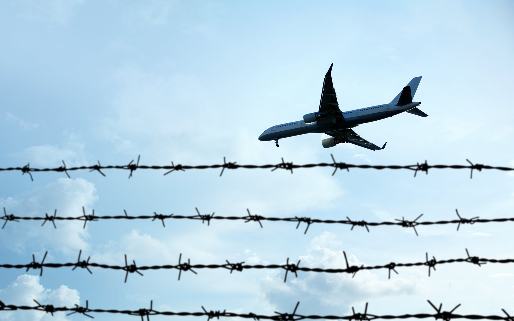 A shot looking up at a plane flying in a blue sky over a barbed wire fence in the foreground.