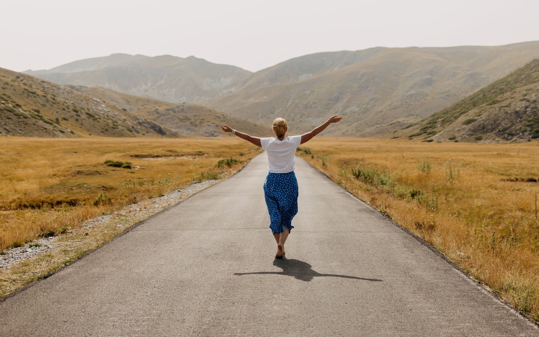 A woman walking alone, shot from behind, on empty mountain road with arms outstretched.