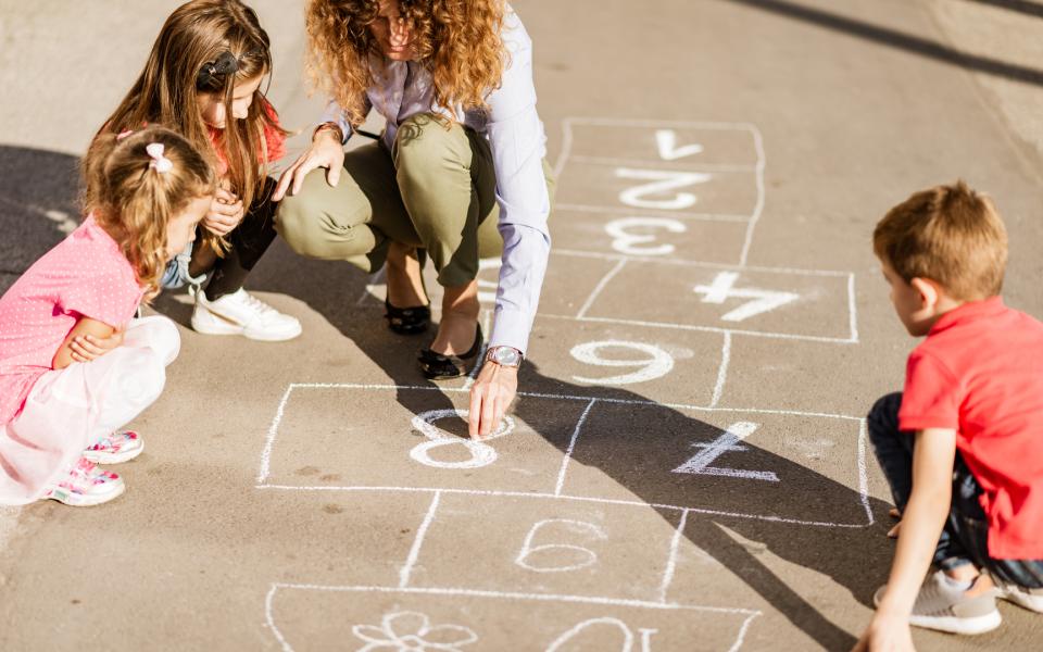Image of a mother and her children drawing a hop scotch board with chalk on the pavement.