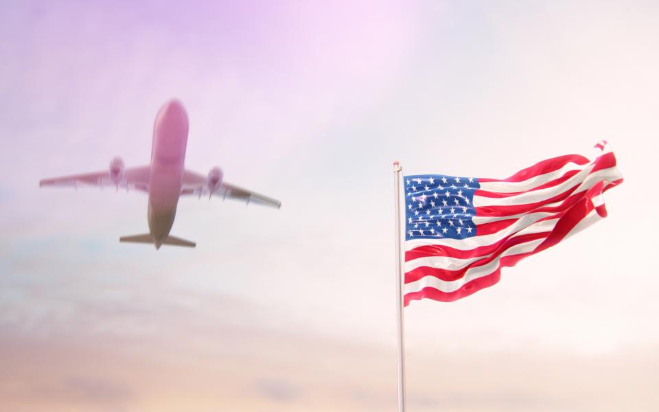 American flag blowing on a pole with an airplane flying above it in the sky.