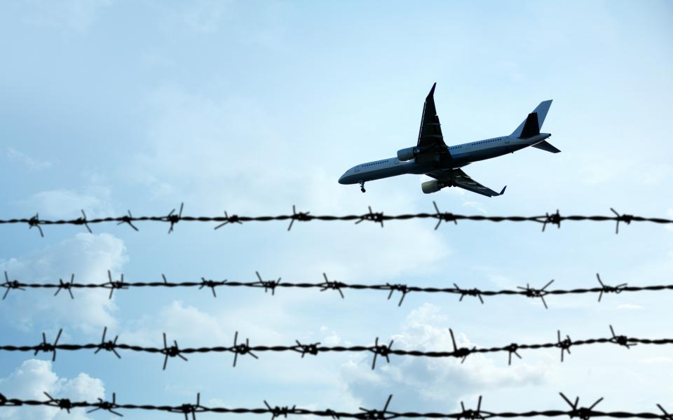 A shot looking up at a plane flying in a blue sky over a barbed wire fence in the foreground.