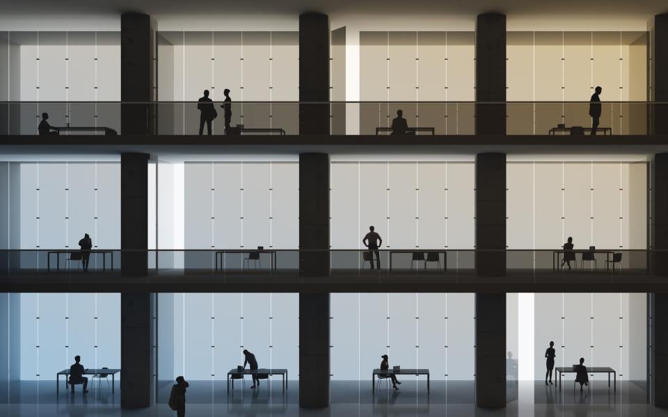 Backlit office workers in their cubicles in an office building of several floors.