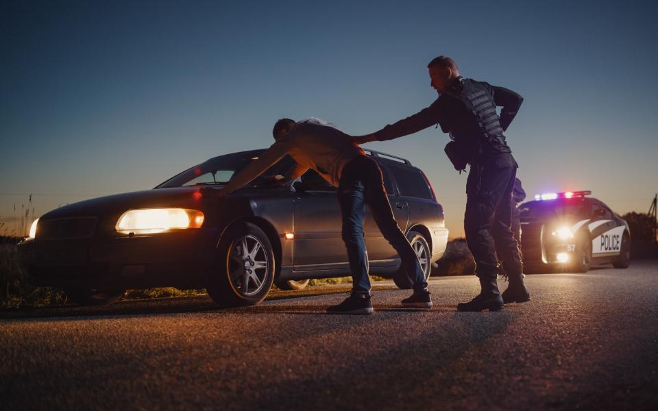 A policeman performing a pat-down search on a fellon who has his hands placed on the hood of the car.