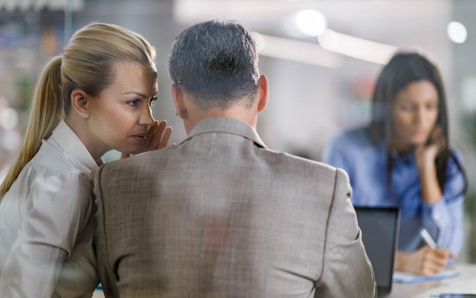 A female coworker whispers into a male coworker's ear during a meeting.